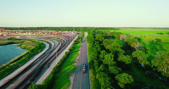 Bird's-eye perspective of a stinger car hauler driving on a highway, parallel to railway tracks, surrounded by vibrant forest and open fields.