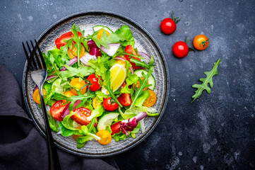 Vegan vegetable salad with tomatoes, cucumber, red onion, lettuce and arugula. Black background, top view