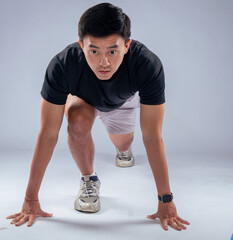 An Asian man in a ready-to-sprint stance, wearing a black t-shirt, gray shorts, and athletic shoes,...