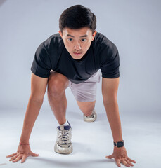 An Asian man in a ready-to-sprint stance, wearing a black t-shirt, gray shorts, and athletic shoes,...