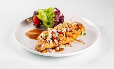 A delectable dish featuring fried fish filet topped with fresh vegetables and nuts, served with a side salad on a white plate.