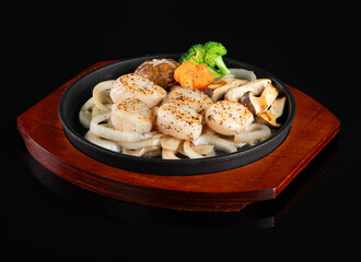 A delicious plate of grilled scallops with noodles, mushrooms, and vegetables on a sizzling hot plate, presented on a wooden tray.