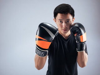 An Asian man in a focused boxing stance, wearing black and orange boxing gloves. He is dressed in a...