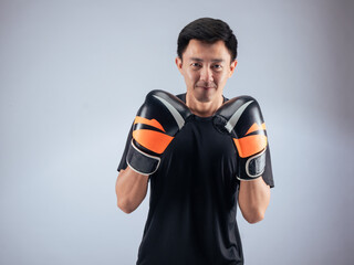 An Asian man in a focused boxing stance, wearing black and orange boxing gloves. He is dressed in a...