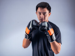 An Asian man in a focused boxing stance, wearing black and orange boxing gloves. He is dressed in a black t-shirt and stands against a white background, ready for action in a fitness studio setting