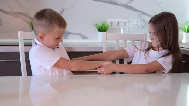Children fight and quarrel over digital tablet. Boy and girl are arguing at computer while sitting at table in living room. Selective focus.