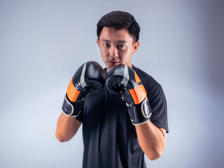 An Asian man in a focused boxing stance, wearing black and orange boxing gloves. He is dressed in a...