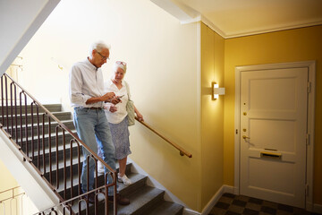 Senior man using smart phone while walking down on stairs with woman in building