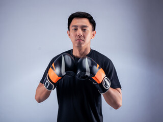 An Asian man in a focused boxing stance, wearing black and orange boxing gloves. He is dressed in a...