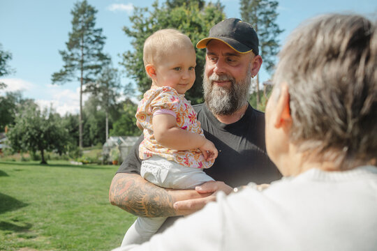 Father carrying baby girl while standing with grandmother in front yard