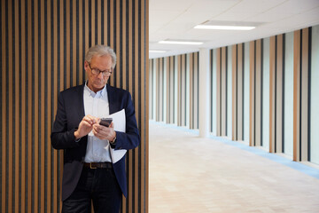 Senior businessman using mobile phone while standing against wall in office