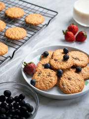 A plate of freshly baked oatmeal raisin cookies garnished with a strawberry, accompanied by a glass of milk and scattered blueberries, set against a light, natural background for a cozy snack scene.