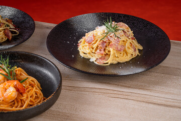 Assorted pasta dishes including a creamy bacon spaghetti, served in black bowls, placed on a wooden table with a red background.