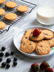 A plate of freshly baked oatmeal raisin cookies garnished with a strawberry, accompanied by a glass of milk and scattered blueberries, set against a light, natural background for a cozy snack scene.