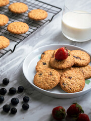 A plate of freshly baked oatmeal raisin cookies garnished with a strawberry, accompanied by a glass of milk and scattered blueberries, set against a light, natural background for a cozy snack scene.