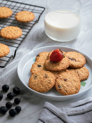 A plate of freshly baked oatmeal raisin cookies garnished with a strawberry, accompanied by a glass of milk and scattered blueberries, set against a light, natural background for a cozy snack scene.