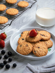 A plate of freshly baked oatmeal raisin cookies garnished with a strawberry, accompanied by a glass of milk and scattered blueberries, set against a light, natural background for a cozy snack scene.