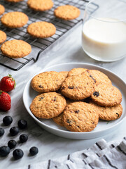 A plate of freshly baked oatmeal raisin cookies garnished with a strawberry, accompanied by a glass of milk and scattered blueberries, set against a light, natural background for a cozy snack scene.