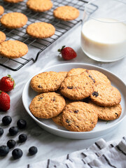 A plate of freshly baked oatmeal raisin cookies garnished with a strawberry, accompanied by a glass of milk and scattered blueberries, set against a light, natural background for a cozy snack scene.