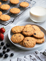 A plate of freshly baked oatmeal raisin cookies garnished with a strawberry, accompanied by a glass of milk and scattered blueberries, set against a light, natural background for a cozy snack scene.