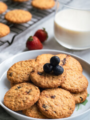 A plate of freshly baked oatmeal raisin cookies garnished with a strawberry, accompanied by a glass of milk and scattered blueberries, set against a light, natural background for a cozy snack scene.