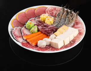 Assorted seafood and meat platter with shrimp, tofu, and vegetables on a white plate over a black background. Perfect for a hotpot meal.