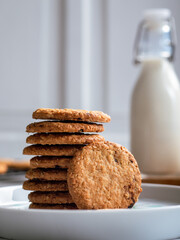 A stack of freshly baked oatmeal raisin cookies neatly arranged on a white plate, with a bottle of milk in the background. The scene is set in a bright kitchen, creating a cozy snack moment.