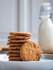 A stack of freshly baked oatmeal raisin cookies neatly arranged on a white plate, with a bottle of milk in the background. The scene is set in a bright kitchen, creating a cozy snack moment.