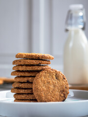 A stack of freshly baked oatmeal raisin cookies neatly arranged on a white plate, with a bottle of milk in the background. The scene is set in a bright kitchen, creating a cozy snack moment.