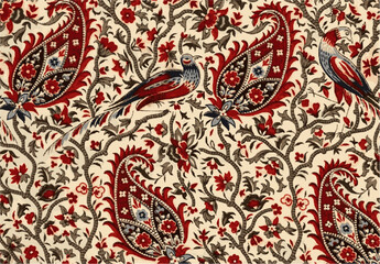 Intricate textile pattern featuring paisley shapes, floral motifs, and exotic birds in warm red, brown, and blue hues.