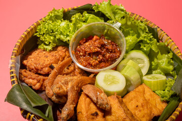 A traditional Indonesian meal served in a woven bamboo basket with fried fish, tofu, cucumber slices, lettuce, and spicy chili paste on banana leaves, creating a vibrant and appetizing presentation