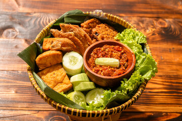 A traditional Indonesian food platter served in a woven bamboo basket, featuring fried fish, tofu, cucumber, lettuce, and sambal chili paste in a wooden bowl, arranged on banana leaves.