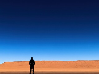 Fototapeta premium A solitary figure stands against a vast desert landscape and clear blue sky, evoking feelings of isolation and contemplation.
