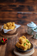 A serving of crispy fried tofu cubes garnished with green chili, presented in a rustic bowl on a wooden table. The background features a blurred bowl of tofu and traditional ceramic containers