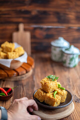 A hand picking up a crispy fried tofu cube from a rustic bowl, served with green chili and...