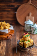 A serving of crispy fried tofu cubes garnished with green chili, presented in a rustic bowl on a wooden table. The background features a blurred bowl of tofu and traditional ceramic containers