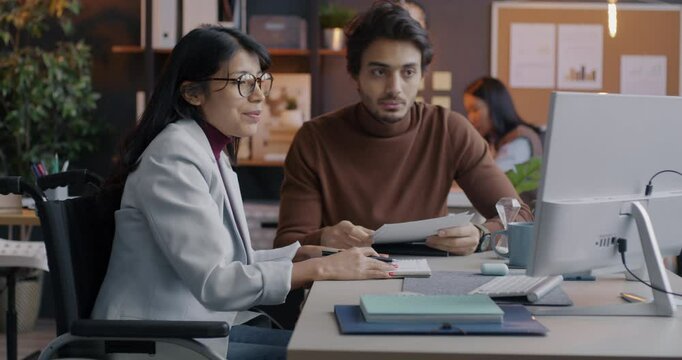 Young woman in wheelchair speaking to male colleague and working with computer in office. Inclusive workspace and successful teamwork concept.