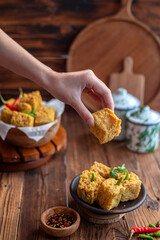 A hand picking up a crispy fried tofu cube from a rustic bowl, served with green chili and...