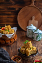 A serving of crispy fried tofu cubes garnished with green chili, presented in a rustic bowl on a wooden table. The background features a blurred bowl of tofu and traditional ceramic containers