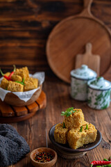 A serving of crispy fried tofu cubes garnished with green chili, presented in a rustic bowl on a wooden table. The background features a blurred bowl of tofu and traditional ceramic containers