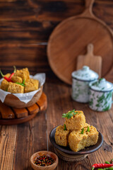 A serving of crispy fried tofu cubes garnished with green chili, presented in a rustic bowl on a wooden table. The background features a blurred bowl of tofu and traditional ceramic containers