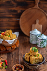 A serving of crispy fried tofu cubes garnished with green chili, presented in a rustic bowl on a wooden table. The background features a blurred bowl of tofu and traditional ceramic containers