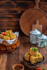 A serving of crispy fried tofu cubes garnished with green chili, presented in a rustic bowl on a wooden table. The background features a blurred bowl of tofu and traditional ceramic containers