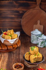 A serving of crispy fried tofu cubes garnished with green chili, presented in a rustic bowl on a wooden table. The background features a blurred bowl of tofu and traditional ceramic containers