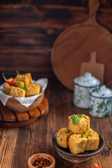 A serving of crispy fried tofu cubes garnished with green chili, presented in a rustic bowl on a wooden table. The background features a blurred bowl of tofu and traditional ceramic containers