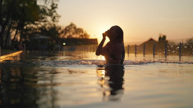 Woman coming up from water and waving wet hair, slow motion, silhouette shot. Carefree lady swimming in beautiful pool in luxury resort in sunset time, sexy lady in swimsuit enjoying summertime trip