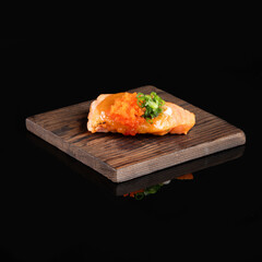 Delicious piece of sushi with salmon, roe, and garnish on a wooden plate, isolated on black background.