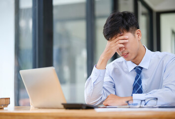 Fototapeta premium Exhausted young man with laptop in office. Stressed and tired young businessman working with laptop. Asian male feel eyes strain.