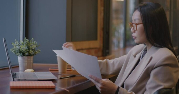 Asian businesswoman rading documents and working with laptop drinking coffee at table in cafe. Business planning and successful person concept.
