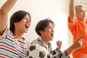 Fototapeta premium Excited Asian Father And Sons Raise Their Hands Showing Win Gesture While Watching Football Match At Home